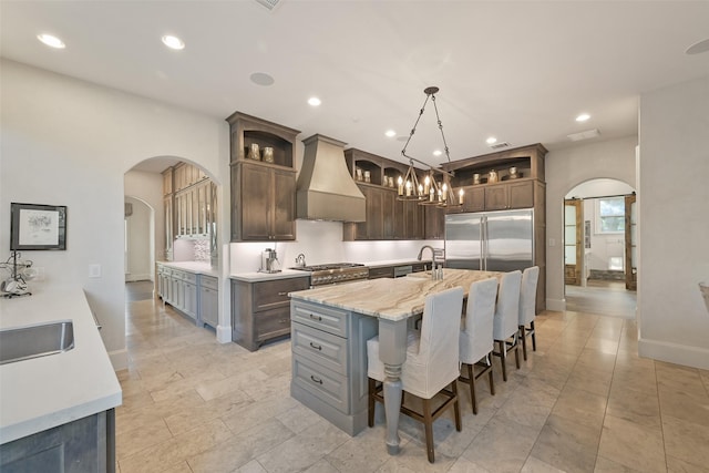 kitchen featuring built in fridge, premium range hood, pendant lighting, a kitchen island with sink, and dark brown cabinets