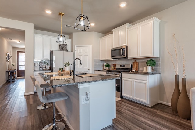 kitchen with pendant lighting, dark stone countertops, white cabinetry, stainless steel appliances, and an island with sink