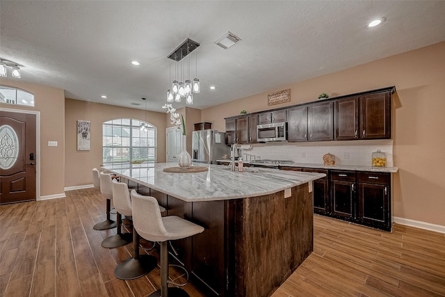 kitchen with pendant lighting, backsplash, dark brown cabinetry, stainless steel appliances, and a center island with sink