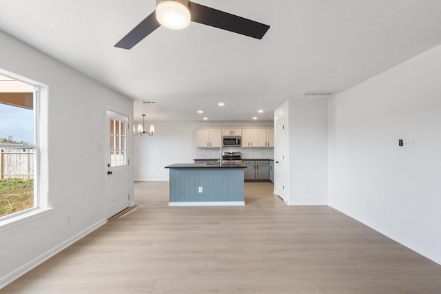 kitchen with appliances with stainless steel finishes, ceiling fan with notable chandelier, pendant lighting, a kitchen island with sink, and light hardwood / wood-style flooring