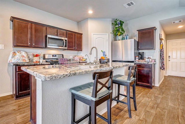 kitchen with sink, stainless steel appliances, light hardwood / wood-style floors, light stone countertops, and a center island with sink