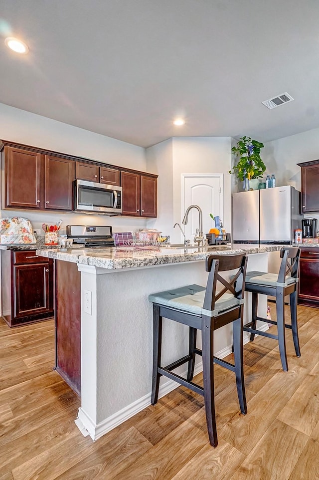 kitchen with stainless steel appliances, an island with sink, light stone countertops, and light wood-type flooring