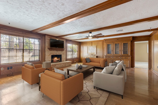 living room featuring wooden walls, a textured ceiling, a brick fireplace, beamed ceiling, and light wood-type flooring