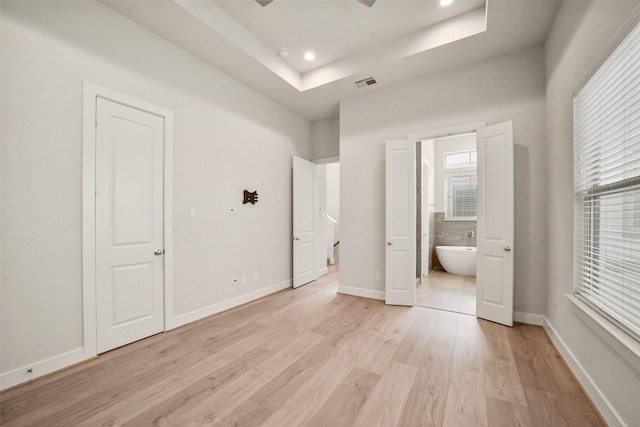 unfurnished bedroom featuring multiple windows, a tray ceiling, light hardwood / wood-style flooring, and ensuite bath