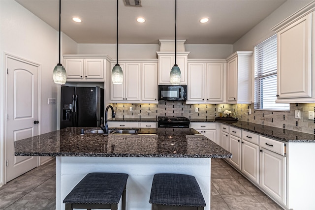 kitchen with sink, a center island with sink, hanging light fixtures, dark stone counters, and black appliances