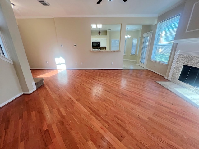 unfurnished living room featuring crown molding, a stone fireplace, light hardwood / wood-style flooring, and ceiling fan with notable chandelier