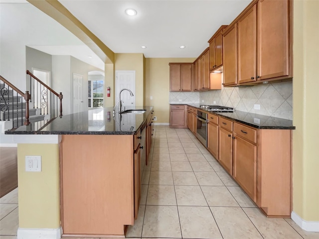 kitchen featuring sink, dark stone countertops, decorative backsplash, light tile patterned floors, and a center island with sink