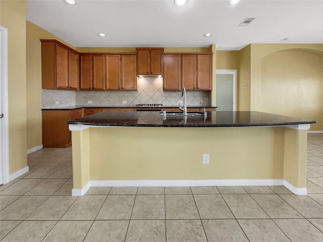 kitchen featuring a kitchen island with sink, sink, stainless steel gas stovetop, and dark stone countertops