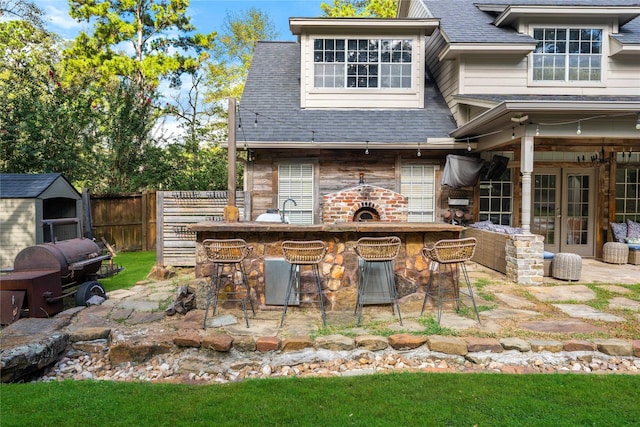 rear view of house featuring a shingled roof, outdoor dry bar, fence, french doors, and a patio area