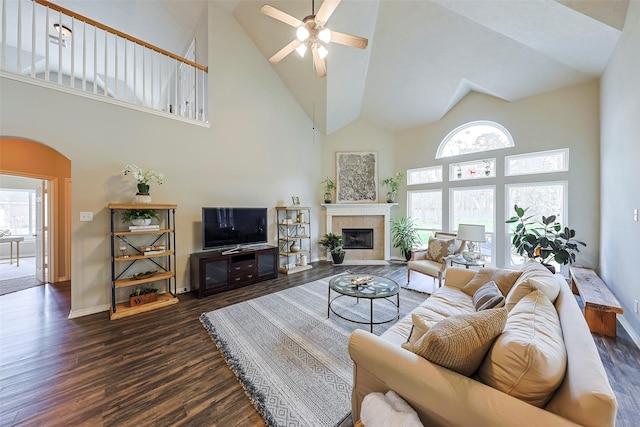 living room with dark wood-type flooring, ceiling fan, a tiled fireplace, and a towering ceiling