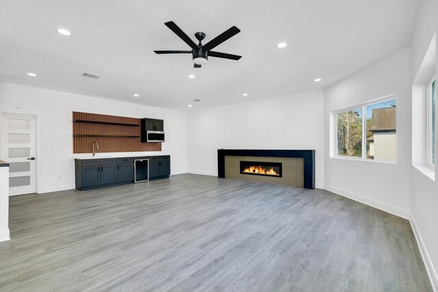 unfurnished living room featuring wine cooler, indoor wet bar, ceiling fan, a tiled fireplace, and light hardwood / wood-style floors