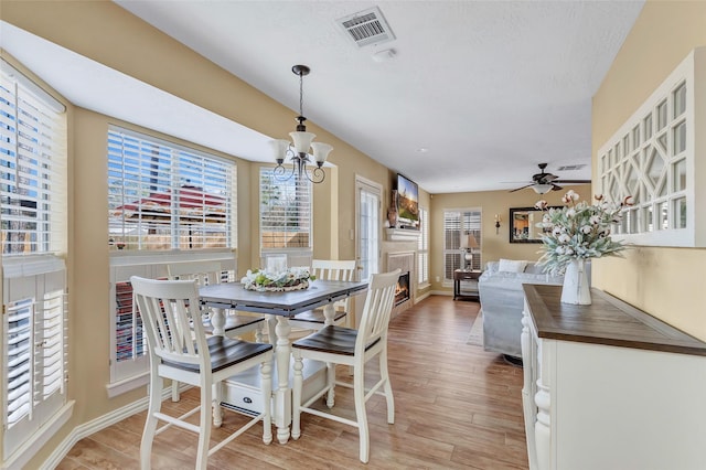 dining area featuring visible vents, baseboards, ceiling fan with notable chandelier, a warm lit fireplace, and light wood-style floors