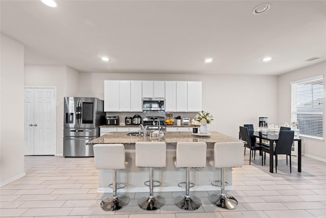 kitchen with a breakfast bar, a kitchen island with sink, white cabinetry, stainless steel appliances, and light stone counters