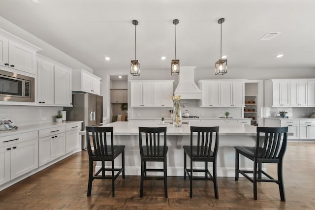 kitchen with a kitchen island with sink, hanging light fixtures, stainless steel appliances, and custom range hood