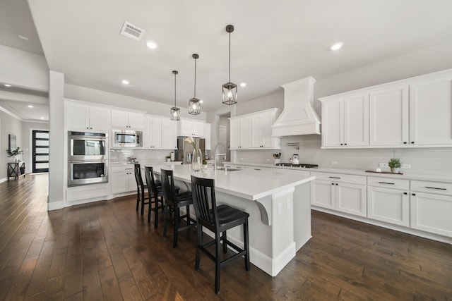 kitchen featuring premium range hood, hanging light fixtures, appliances with stainless steel finishes, an island with sink, and white cabinets