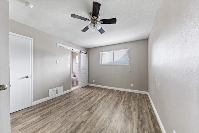 unfurnished bedroom with ensuite bath, wood-type flooring, a textured ceiling, ceiling fan, and a barn door