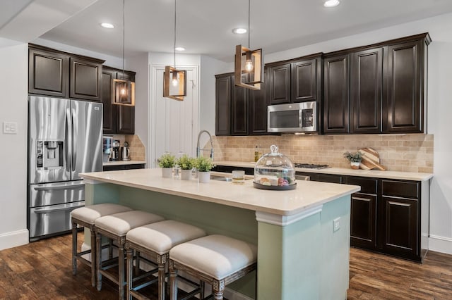 kitchen with dark brown cabinets, hanging light fixtures, dark hardwood / wood-style flooring, stainless steel appliances, and a kitchen island with sink