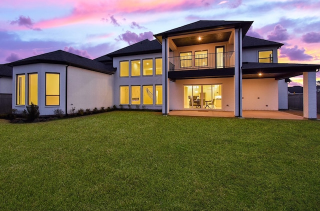 back house at dusk with a lawn, a patio, and a balcony