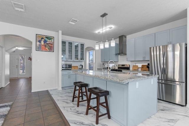 kitchen featuring decorative light fixtures, a kitchen island with sink, stainless steel appliances, light stone countertops, and wall chimney exhaust hood