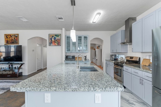 kitchen featuring sink, pendant lighting, stainless steel appliances, a kitchen island with sink, and wall chimney range hood