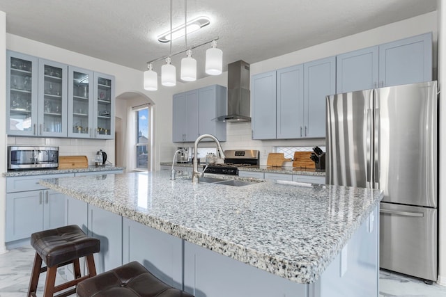 kitchen with wall chimney range hood, sink, hanging light fixtures, stainless steel appliances, and light stone counters