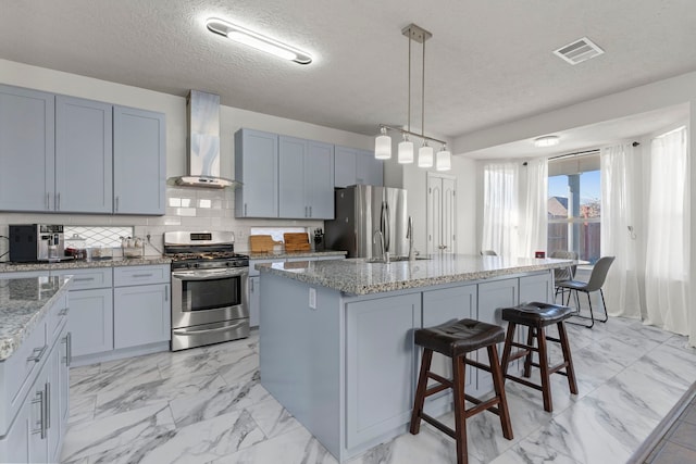 kitchen with wall chimney exhaust hood, light stone counters, decorative light fixtures, a center island with sink, and stainless steel appliances