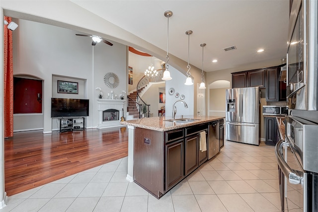 kitchen with appliances with stainless steel finishes, decorative light fixtures, light stone counters, dark brown cabinets, and a center island with sink