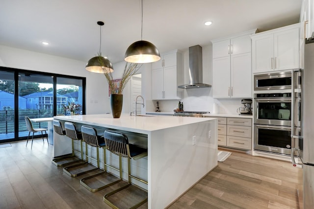 kitchen with pendant lighting, white cabinetry, sink, a kitchen island with sink, and wall chimney range hood