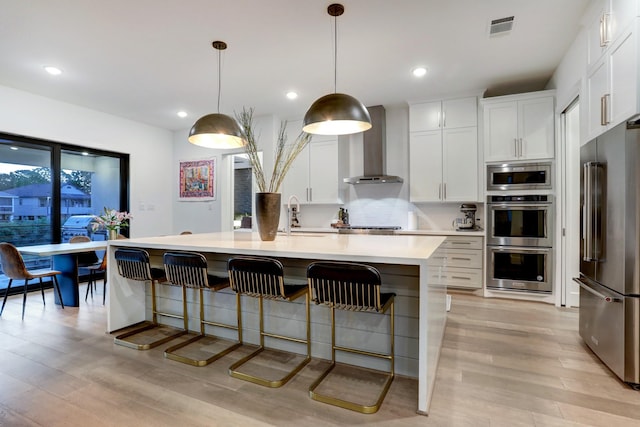 kitchen featuring white cabinetry, appliances with stainless steel finishes, an island with sink, pendant lighting, and wall chimney range hood