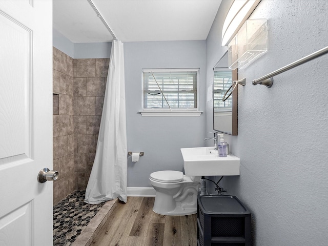 bathroom featuring toilet, a sink, wood finished floors, tiled shower, and baseboards