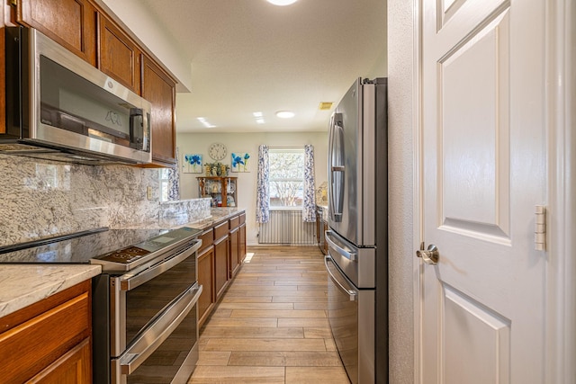 kitchen featuring stainless steel appliances, decorative backsplash, brown cabinetry, light stone countertops, and light wood-type flooring