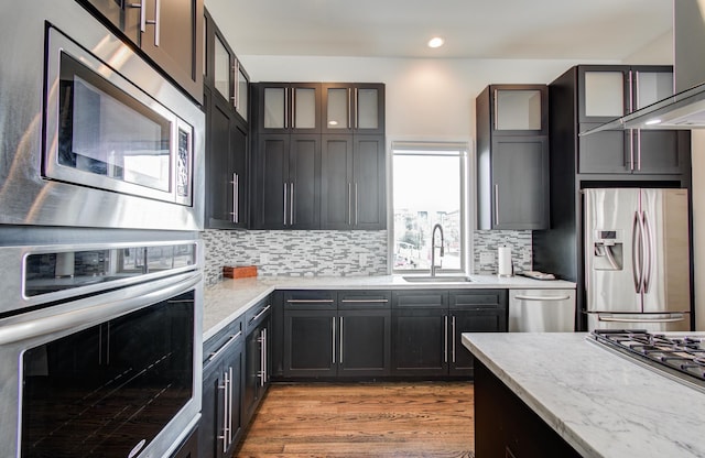 kitchen with a sink, wall chimney range hood, appliances with stainless steel finishes, decorative backsplash, and glass insert cabinets