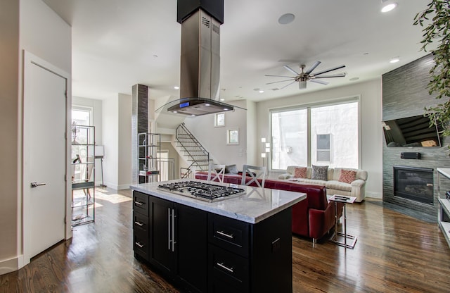 kitchen featuring island exhaust hood, stainless steel gas stovetop, open floor plan, a large fireplace, and dark cabinetry