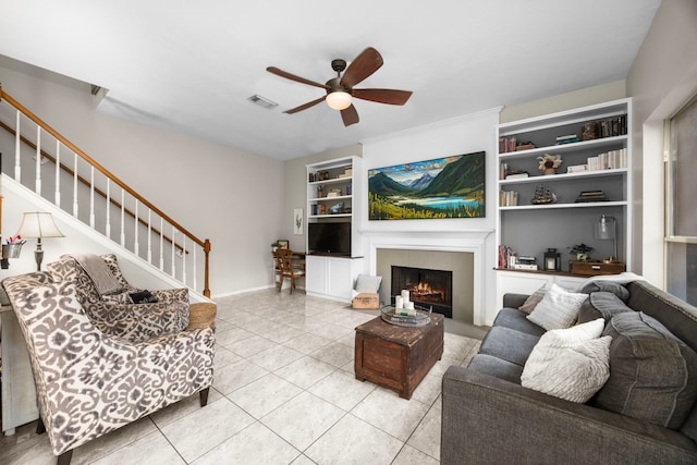 tiled living room with a tiled fireplace, built in features, and ceiling fan