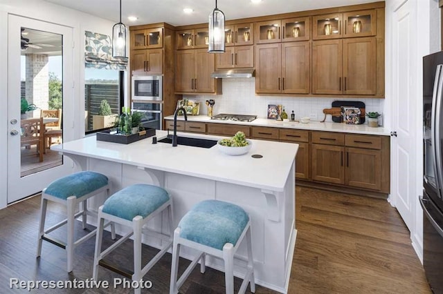 kitchen with under cabinet range hood, brown cabinets, dark wood finished floors, stainless steel appliances, and a sink