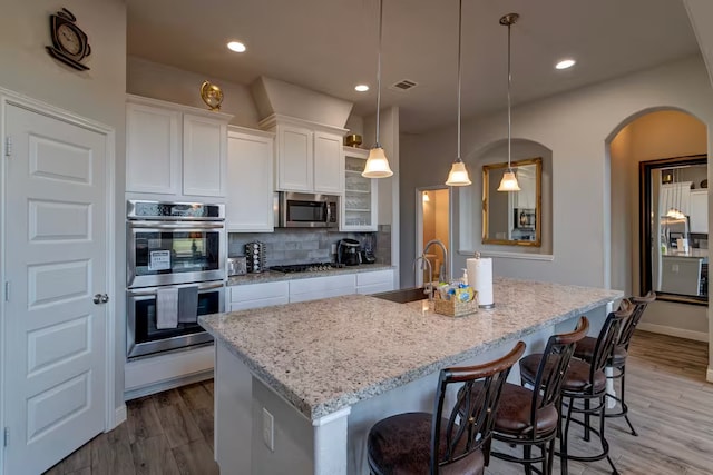 kitchen featuring appliances with stainless steel finishes, white cabinets, hanging light fixtures, light stone countertops, and a center island with sink