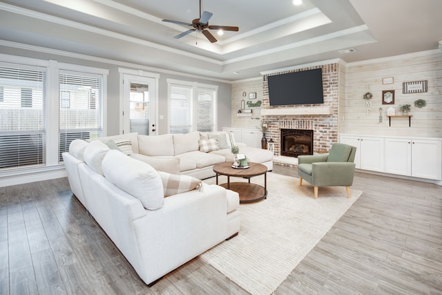 living room with a brick fireplace, light hardwood / wood-style flooring, ornamental molding, a tray ceiling, and ceiling fan