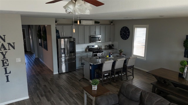 kitchen featuring gray cabinets, appliances with stainless steel finishes, dark stone counters, dark wood-type flooring, and a center island with sink