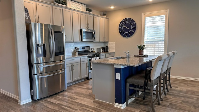 kitchen with sink, appliances with stainless steel finishes, gray cabinets, light stone countertops, and a kitchen island with sink