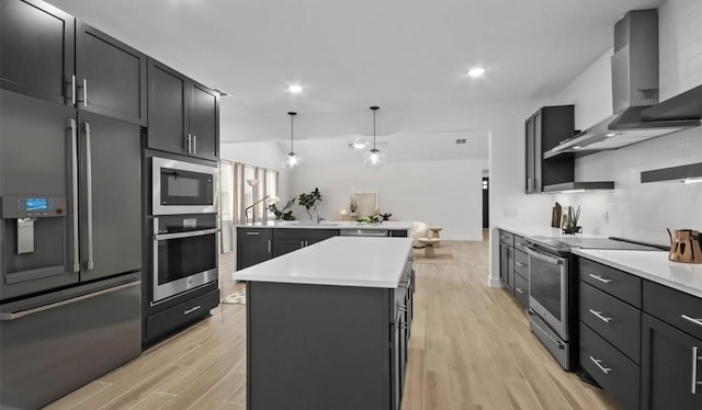 kitchen with wall chimney exhaust hood, hanging light fixtures, light wood-type flooring, appliances with stainless steel finishes, and a kitchen island