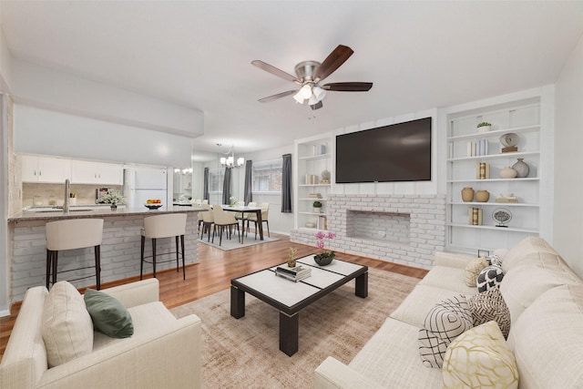 living room with ceiling fan with notable chandelier, sink, light hardwood / wood-style floors, and a brick fireplace