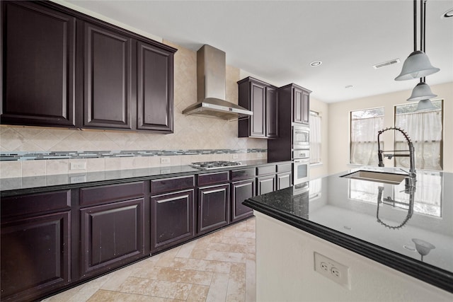 kitchen with a sink, hanging light fixtures, appliances with stainless steel finishes, backsplash, and wall chimney exhaust hood