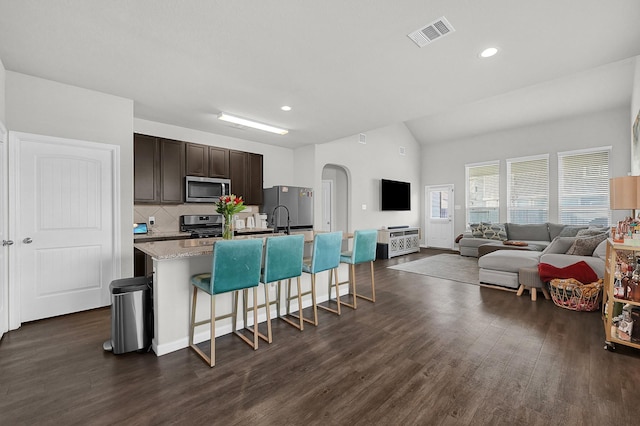 kitchen featuring appliances with stainless steel finishes, a breakfast bar, light stone counters, dark brown cabinets, and a center island with sink