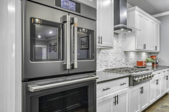 kitchen featuring wall chimney range hood, appliances with stainless steel finishes, ornamental molding, white cabinets, and decorative backsplash