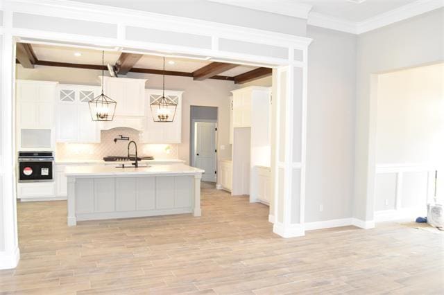 kitchen with beamed ceiling, white cabinetry, an island with sink, hanging light fixtures, and stainless steel oven