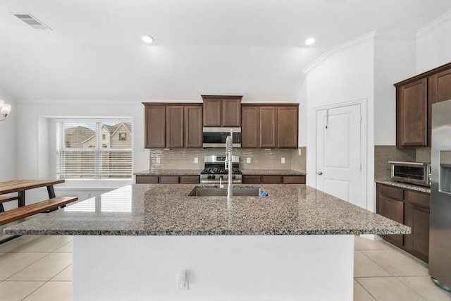 kitchen with a kitchen island with sink, light tile patterned floors, stainless steel appliances, and dark stone countertops