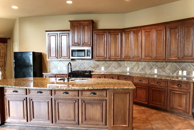 kitchen with sink, black fridge, tasteful backsplash, light stone counters, and a center island with sink