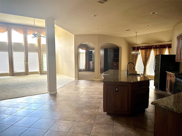 kitchen with sink, a kitchen island with sink, hanging light fixtures, black appliances, and dark stone counters