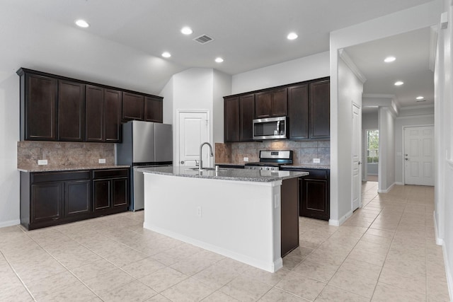 kitchen featuring sink, dark brown cabinets, stainless steel appliances, light stone counters, and an island with sink