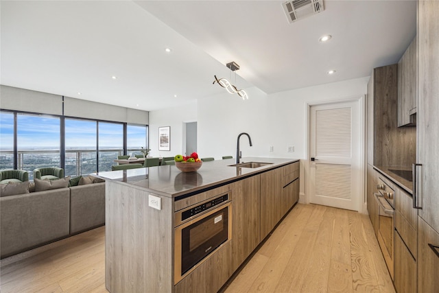 kitchen with sink, stainless steel oven, hanging light fixtures, a kitchen island with sink, and light hardwood / wood-style floors